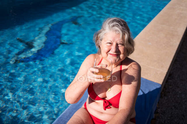 Alto ângulo de turista sênior encantado em biquíni rindo brilhantemente enquanto arrefece na beira da piscina com bebida — Fotografia de Stock
