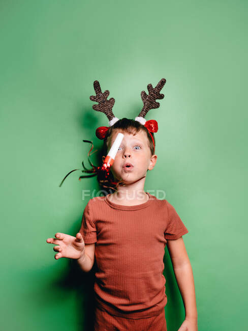 Menino vestindo chifres de rena headband e festa festiva soprador de pé contra fundo verde e olhando para longe — Fotografia de Stock