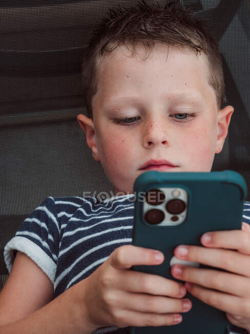 Serious boy in t shirt lying alone and browsing Internet on modern mobile phone — Stock Photo