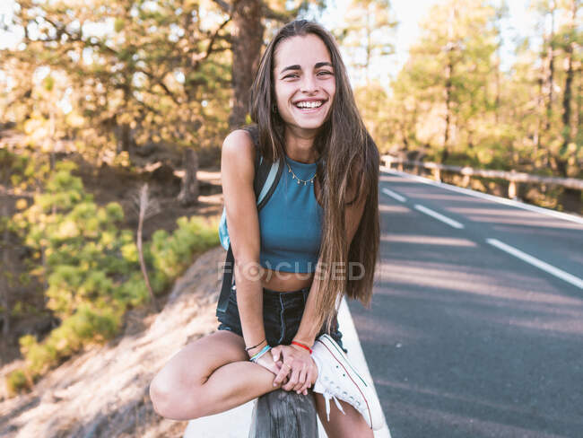 Sonriente adolescente en gumshoe de moda tocando el antebrazo mientras mira la cámara en la valla en Tenerife España - foto de stock
