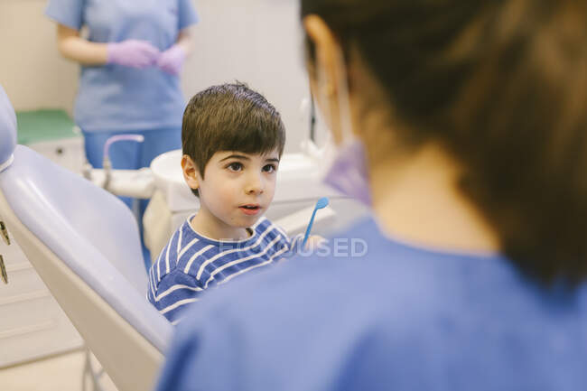 Chico curioso con instrumento dental hablando con el médico de la cosecha en la máscara durante la cita en el consultorio dental moderno - foto de stock