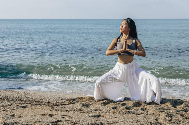 Jeune femme asiatique rêveuse avec des mains priantes accroupi dans la pose Utkata Konasana tout en pratiquant le yoga et en regardant loin sur le rivage de la mer — Photo de stock