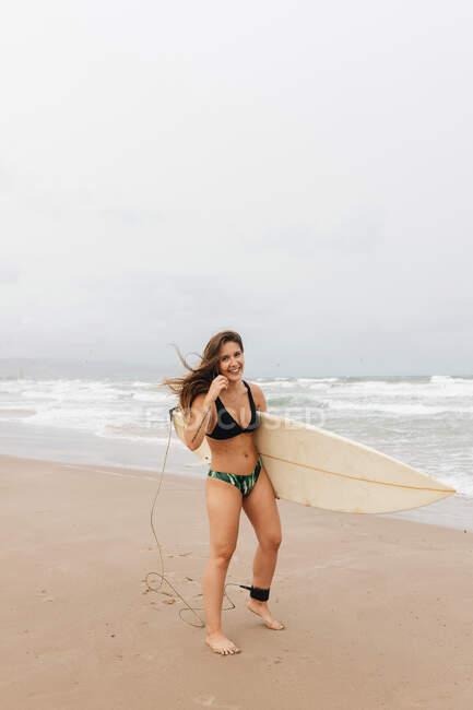 Joven deportista alegre en traje de baño con tabla de surf mirando a la cámara en la costa arenosa contra el océano tormentoso - foto de stock