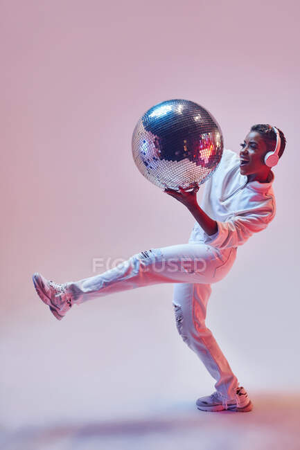 Mujer étnica fresca con pelo corto en auriculares inalámbricos y ropa de moda bailando hip hop con la boca abierta mirando hacia otro lado mientras sostiene la bola de brillo sobre fondo violeta - foto de stock