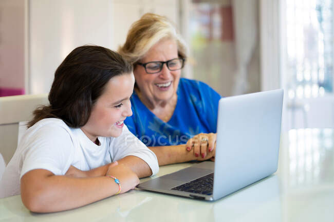 Petite-fille et grand-mère souriantes assises à table et utilisant un ordinateur portable dans la pièce lumineuse de l'appartement — Photo de stock