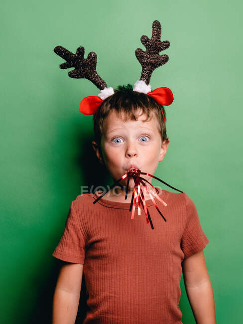 Junge mit Rentierhörnern Stirnband und Festtagsgebläse im Mund steht vor grünem Hintergrund und blickt in die Kamera — Stockfoto