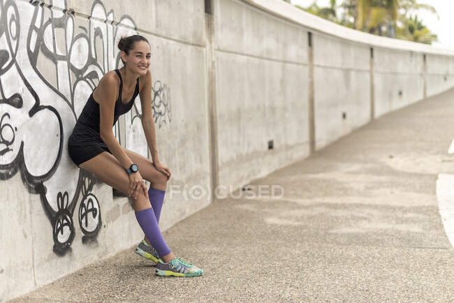 Seitenansicht einer positiven athletischen Läuferin in Sportbekleidung, die sich während einer Trainingspause in der Stadt an die Wand lehnt — Stockfoto