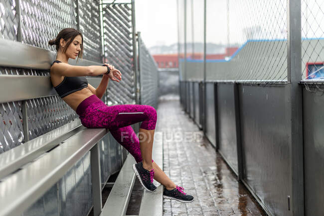 Side view full body of fit female sitting on bench and looking at wearable tracker during fitness training — Stock Photo