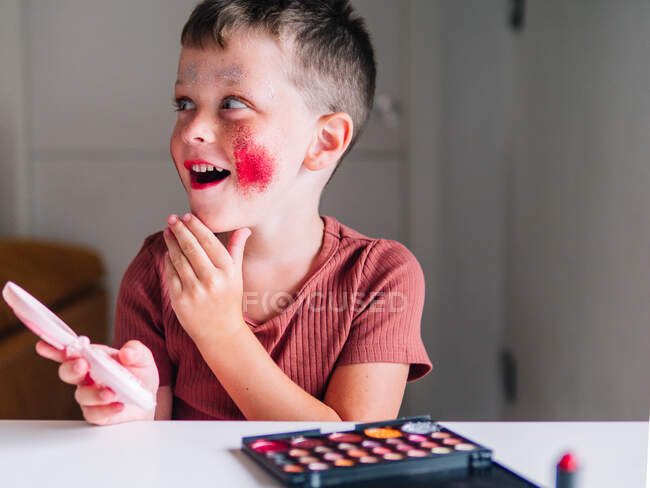 Criança incrível com rosto sujo e espelho cobrindo a boca enquanto olha para a mesa com paleta de sombra na casa — Fotografia de Stock