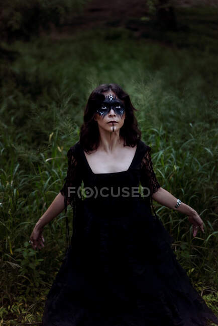 Зверху містична відьма в довгій чорній сукні і з пофарбованим обличчям, що стоїть в темному похмурому лісі — стокове фото