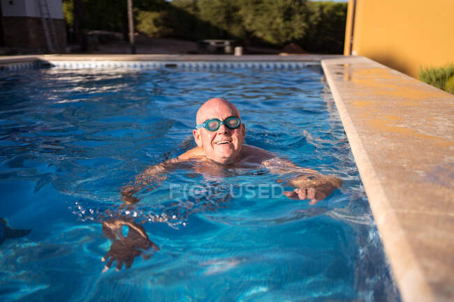 Entspannter Senior in Brille schwimmt im transparenten sauberen Poolwasser und entspannt sich an heißen Sommertagen — Stockfoto