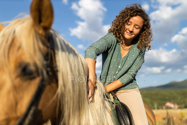 Smiling female with curly hair caressing stallion mane under cloudy blue sky in sunlight — Stock Photo