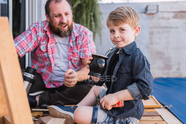 Criança sincera com cabaça de calabash de bebida infundida contra pai barbudo alegre com termo no fundo embaçado — Fotografia de Stock