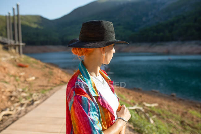 Mujer vista lateral en un lago, mirando hacia otro lado con una mano sosteniendo un sombrero negro - foto de stock