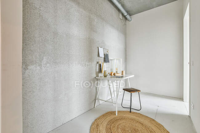 Decoración en la mesa contra el taburete y la alfombra en el suelo bajo el tubo en la pared gris en el paso - foto de stock