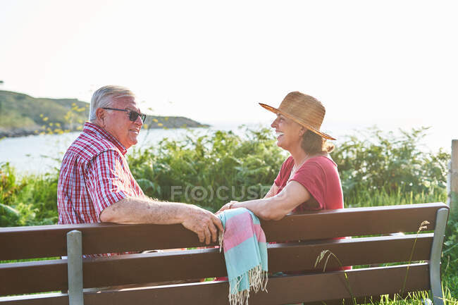 Side view of senior couple sitting looking at each other on wooden bench and enjoying summer day on shore of pond — Stock Photo