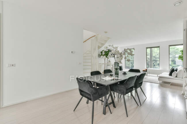 Interior de la zona de comedor con mesa grande con ramo de flores y sillas en apartamento moderno en el día y en el fondo la sala de estar y ventanas luminosas. - foto de stock