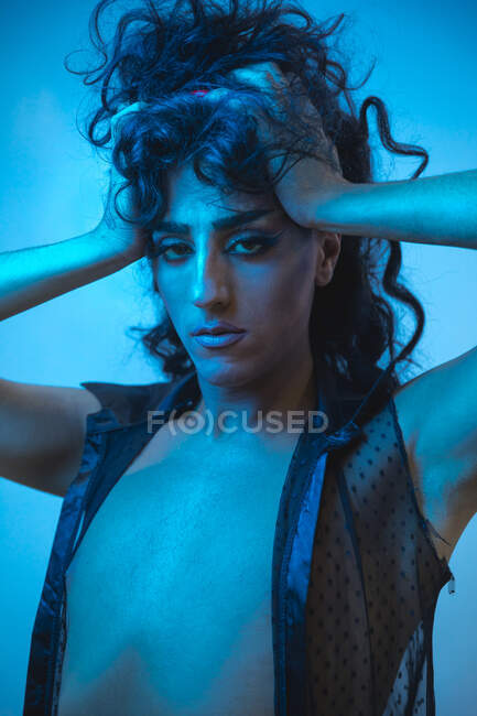 Joven transexual modelo masculino con maquillaje en chaqueta elegante mirando a la cámara sobre fondo azul - foto de stock
