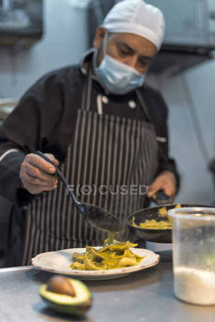 Aufmerksame reife ethnische männliche Köchin in steriler Maske gießt Avocadosauce auf gekochte Pasta bei der Arbeit im Restaurant — Stockfoto