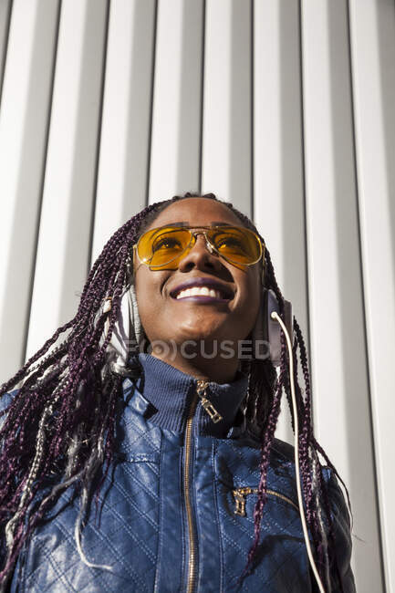 D'en bas jeune femme afro-américaine heureuse avec des tresses afro vêtues d'une veste bleue et des lunettes de soleil élégantes profitant de la musique à travers des écouteurs tout en se refroidissant au soleil contre un mur rayé — Photo de stock