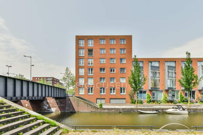 Canale del fiume sotto il ponte situato vicino edificio in mattoni residenziali in città sotto cielo blu senza nuvole nella giornata di sole — Foto stock
