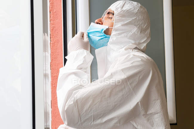 Vista lateral del médico varón adulto en equipo de protección personal con los ojos cerrados respirando aire fresco en el hospital - foto de stock