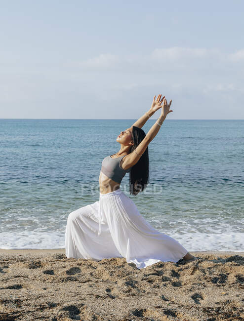 Вид сбоку этнической женщины, откинувшейся назад, стоя в позе Ашты Чандрасаны во время занятий йогой на песчаном побережье против океана — стоковое фото