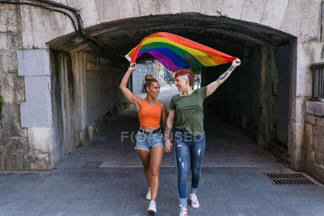 Cheerful cool young homosexual girlfriends with LGBTQ flag holding hands while looking at each other and strolling on urban pavement — Stock Photo