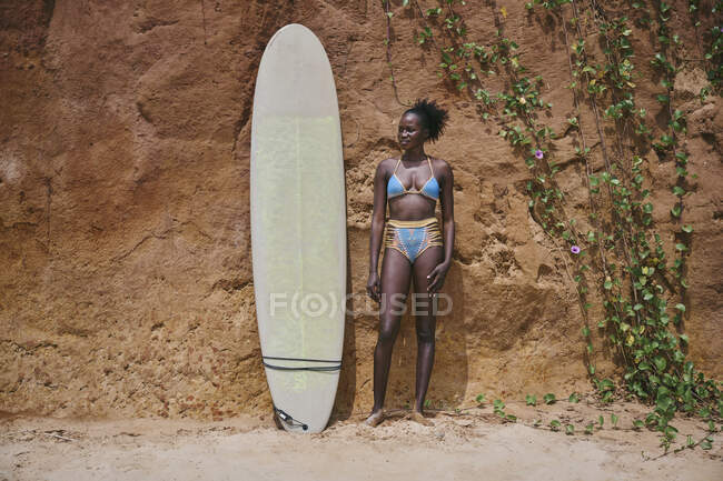 Front view of African American female athlete looking away with surfboard from an area of the beach and in front of a clay rock with plants on its side — Stock Photo