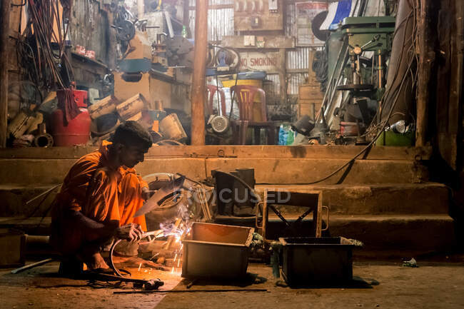 ÍNDIA, BANGLADESH - DEZEMBRO 6, 2015: Vista lateral do mecânico masculino em vestuário de trabalho e luvas de pé agachado trabalhando com fábrica de fundição de metal — Fotografia de Stock