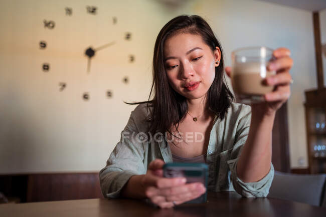 Young ethnic female with glass of coffee surfing internet on cellphone at table in house room — Stock Photo