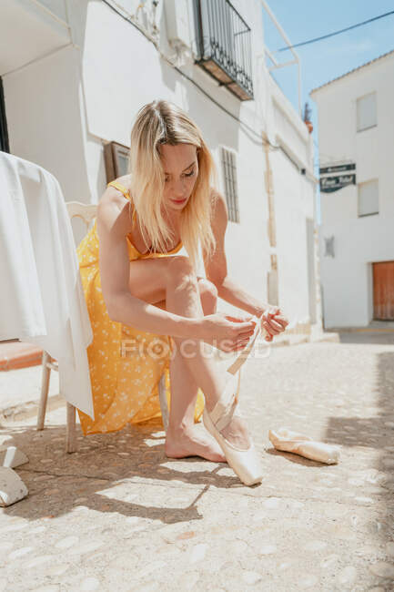 Full body of ballerina in dress sitting on chair and putting on pointe shoes in paved alley — Stock Photo
