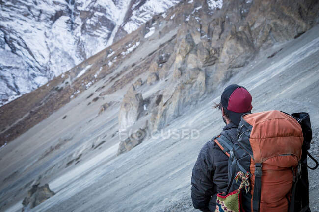 Vista trasera del explorador irreconocible con la mochila de pie en la empinada ladera rocosa de la montaña mientras viaja por las tierras altas de Nepal - foto de stock