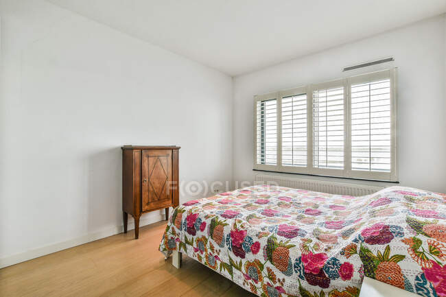 Funda de cama con adorno floral contra cómoda vintage y ventana con jalousie en casa ligera - foto de stock