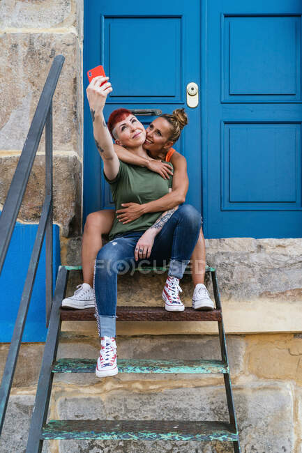 Улыбающиеся гомосексуальные женщины с татуировками, делающие автопортрет по мобильному телефону у входной двери в городе — стоковое фото
