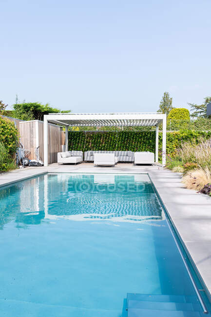 Eau bleue claire dans la piscine située près de la zone lounge et plantes vertes sous un ciel bleu sans nuages au soleil — Photo de stock