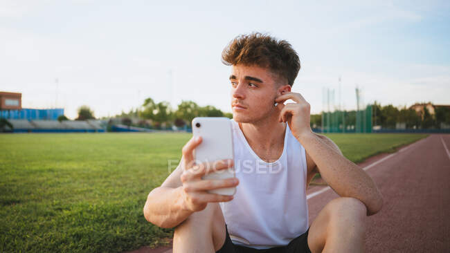 Giovane atleta maschile contemplativo in maglietta con taglio di capelli moderno che ascolta musica da auricolare wireless mentre guarda lontano nello stadio — Foto stock