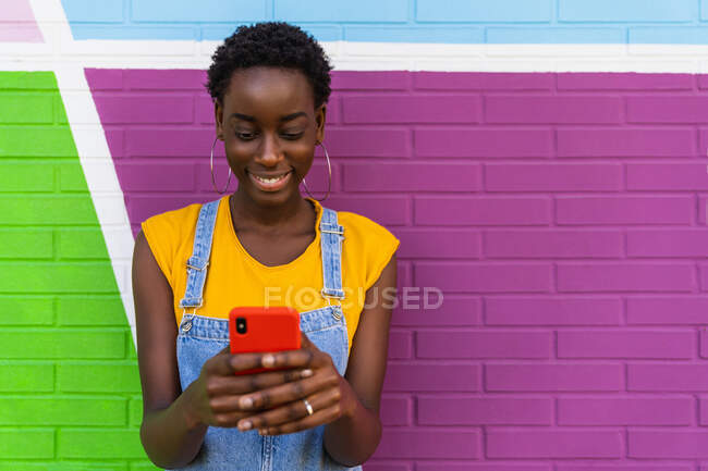 Mujer afroamericana en overoles de mezclilla de pie cerca de la pared colorida y el teléfono celular de navegación - foto de stock