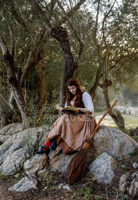 Focada bruxa fêmea leitura livro mágico enquanto sentado na rocha perto de vassoura em bosques — Fotografia de Stock