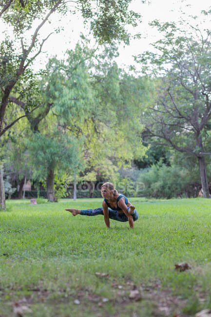 Окаменелая женщина в стильной одежде, делающая позу Светлячка, практикуя йогу в парке в дневное время — стоковое фото