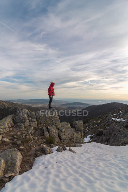 Vista lateral de cuerpo completo del irreconocible viajero masculino en ropa de abrigo parado en el borde rocoso y admirando el paisaje en la nevada Sierra de Guadarrama - foto de stock