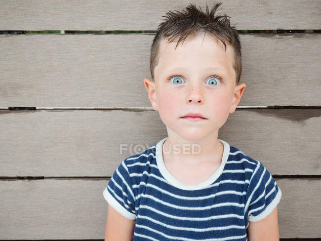 Удивительный мальчик с мокрыми волосами в полосатой футболке смотрит в камеру с шокированным выражением лица на деревянной стене — стоковое фото