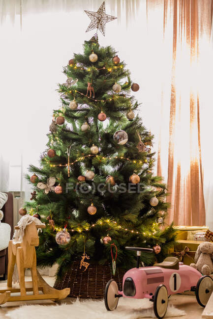 Wooden rocking horse and velomobile under Christmas tree decorated with fairy lights and baubles placed near window — Stock Photo