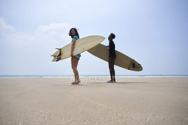 Back view of young multiethnic female surfers with surfboards speaking while strolling on sandy shore and looking at camera — Stock Photo