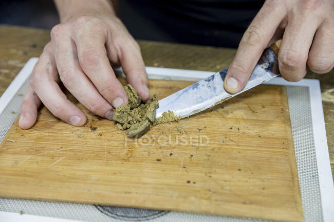 Врожай невизначений чоловік з ножем, що розмелює сушені шматочки рослини канабісу на дерев'яній дошці в робочому просторі — стокове фото