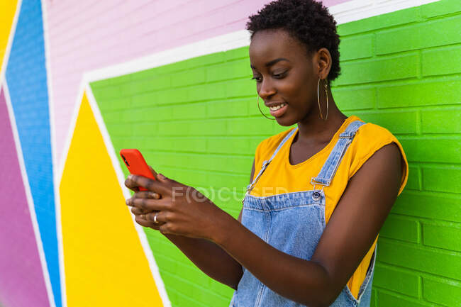 African American female in denim overalls standing near colorful wall and browsing cellphone — Stock Photo