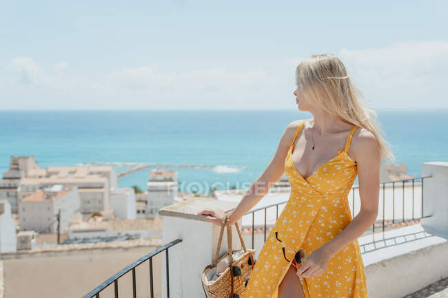Female traveler in dress standing near fence and admiring old coastal city and blue sea during summer vacation — Stock Photo