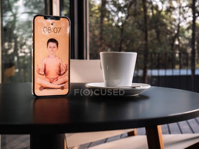 Зображення дитини, що робить йогу в позі Лотоса на екрані мобільного телефону, розміщеного на столі біля чашки напою — стокове фото