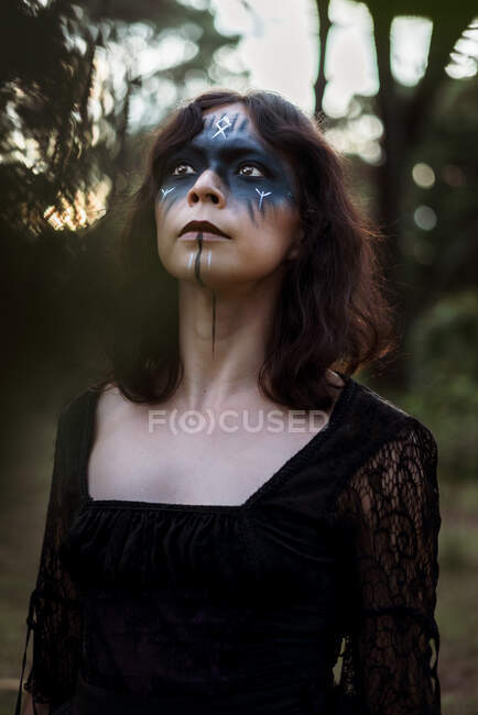 Bruja mística en vestido negro largo y con la cara pintada de pie mirando hacia arriba en los bosques oscuros y sombríos - foto de stock