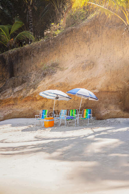 Vista paisagem de poltronas e geladeira portátil sob guarda-sóis na costa arenosa contra montagem à luz do sol — Fotografia de Stock